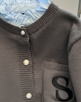 Nahaufnahme der grauen Strickjacke mit strukturiertem Stoff, glänzenden Knöpfen und sauber eingearbeitetem „S“-Patch.