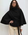Model trägt ein schwarzes Cape-Oberteil mit weitem Schnitt und Bindegürtel, kombiniert mit heller Hose im Studio.
