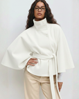 Weißes Cape-Oberteil mit fließender Silhouette und Gürtel, getragen vor neutralem Hintergrund.