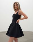 Model trägt ein schwarzes Kleid mit ausgestelltem Ballonrock und klarer Silhouette in einem sonnigen Innenraum.