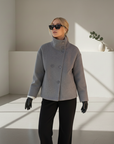 Model trägt eine graue Wolljacke mit lockerer Passform und modernem Schnitt in einem minimalistischen Innenraum.