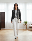 Model trägt eine schwarze Lederjacke und weiße Hose in eleganter Pose innerhalb eines hellen Innenraums.