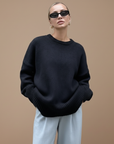 Model trägt einen schwarzen Oversize-Pullover mit weiter Passform und weichem Strickmaterial in einem hellen Innenraum.