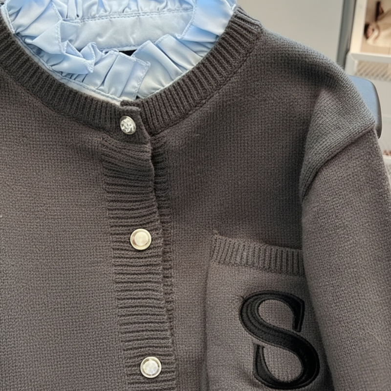 Nahaufnahme der grauen Strickjacke mit strukturiertem Stoff, glänzenden Knöpfen und sauber eingearbeitetem „S“-Patch.
