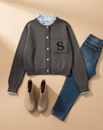 Graue Strickjacke mit Letter-Patch flach ausgelegt, kombiniert mit Jeans und Accessoires auf hellem Untergrund.