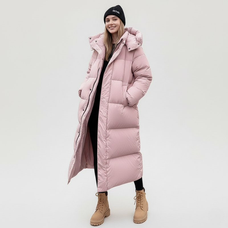 Model trägt einen rosafarbenen, langen Damen-Daunenmantel mit Kapuze, kombiniert mit Stiefeln im Studio.