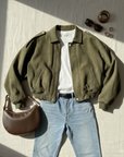 Olivgrüne Damenjacke mit Jeans, Tasche und Accessoires als Flatlay auf hellem Untergrund arrangiert.
