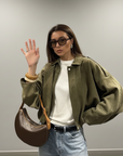 Model trägt eine olivgrüne Jacke mit Sonnenbrille und Jeans in einem lässigen, modernen Look.