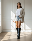Model trägt kniehohe schwarze Stiefel aus glattem Material, kombiniert mit Shorts und Cardigan in einer hellen Innenraumszene.