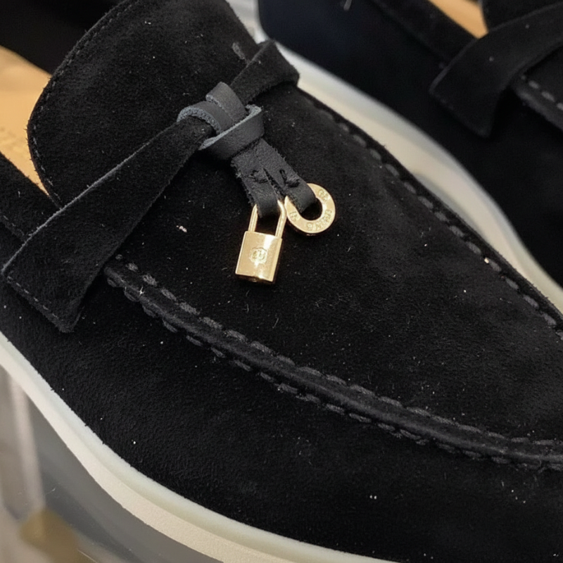 Nahaufnahme eines schwarzen Loafers mit goldener Verschlussverzierung und weicher Wildlederstruktur.