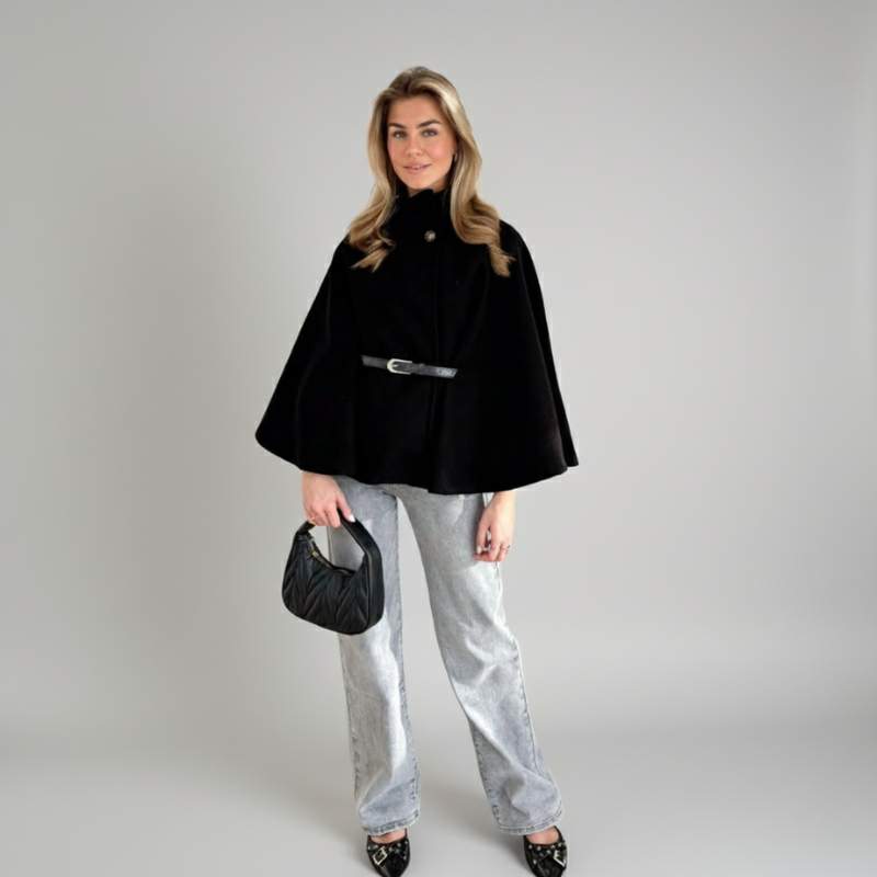 Model trägt einen schwarzen Damenponcho mit Taillengürtel, kombiniert mit heller Hose und Handtasche im Studio.