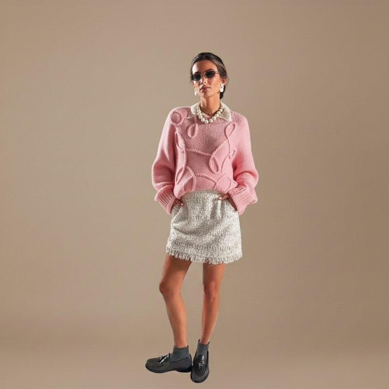 Rosa Pullover mit strukturiertem Strick, kombiniert mit Rock und Accessoires in einer hellen Innenraumszene.