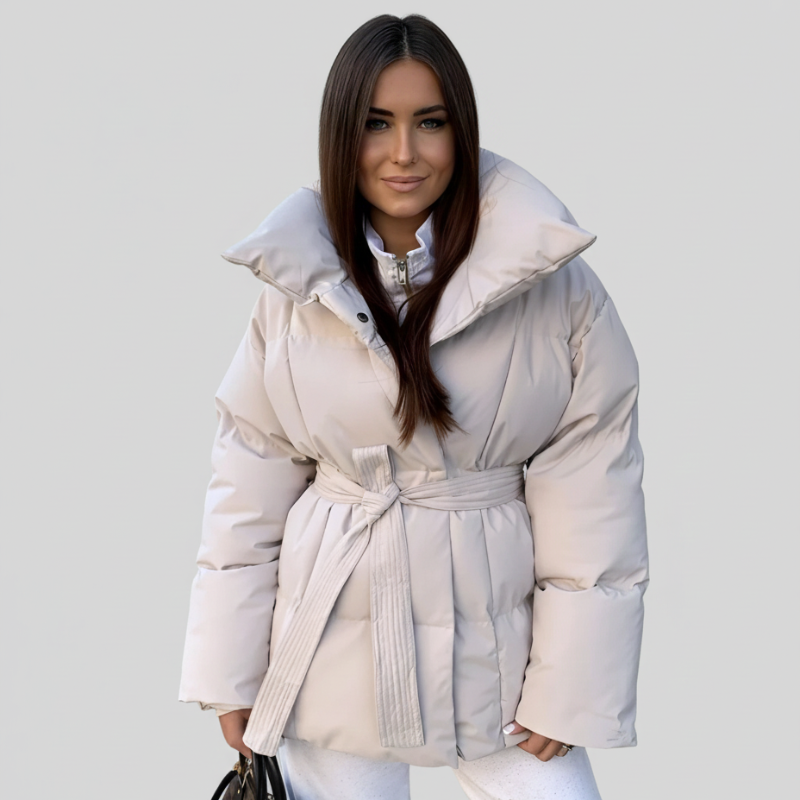 Frau trägt eine helle Steppjacke mit Gürtel, kombiniert mit heller Hose in einem lichtdurchfluteten Innenraum.