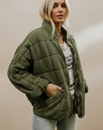 Model trägt eine olivgrüne Steppjacke mit lockerer Passform und funktionalen Details in einem hellen Innenraum.