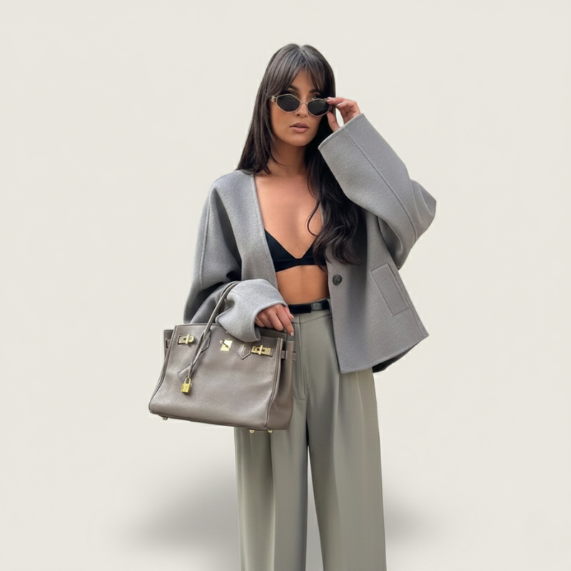 Modell zeigt eine graue, kastige Jacke kombiniert mit weiter Hose und Handtasche im neutralen Studio.