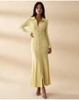 Model zeigt ein gelbes langes Strickkleid mit strukturiertem Strickmuster und Knöpfen in weichem Indoor-Licht.