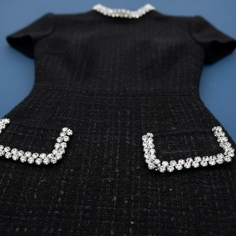 Nahaufnahme des schwarzen Kleids mit strukturierter Oberfläche und verzierten Taschen mit kontrastierender Kantenverarbeitung.