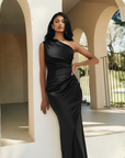 Model trägt ein schwarzes One-Shoulder-Abendkleid mit drapiertem Detail und fließendem Stoff in heller Umgebung.